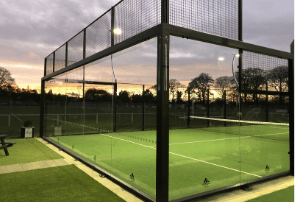 Completed glass-and-mesh padel court with floodlit synthetic turf at dusk